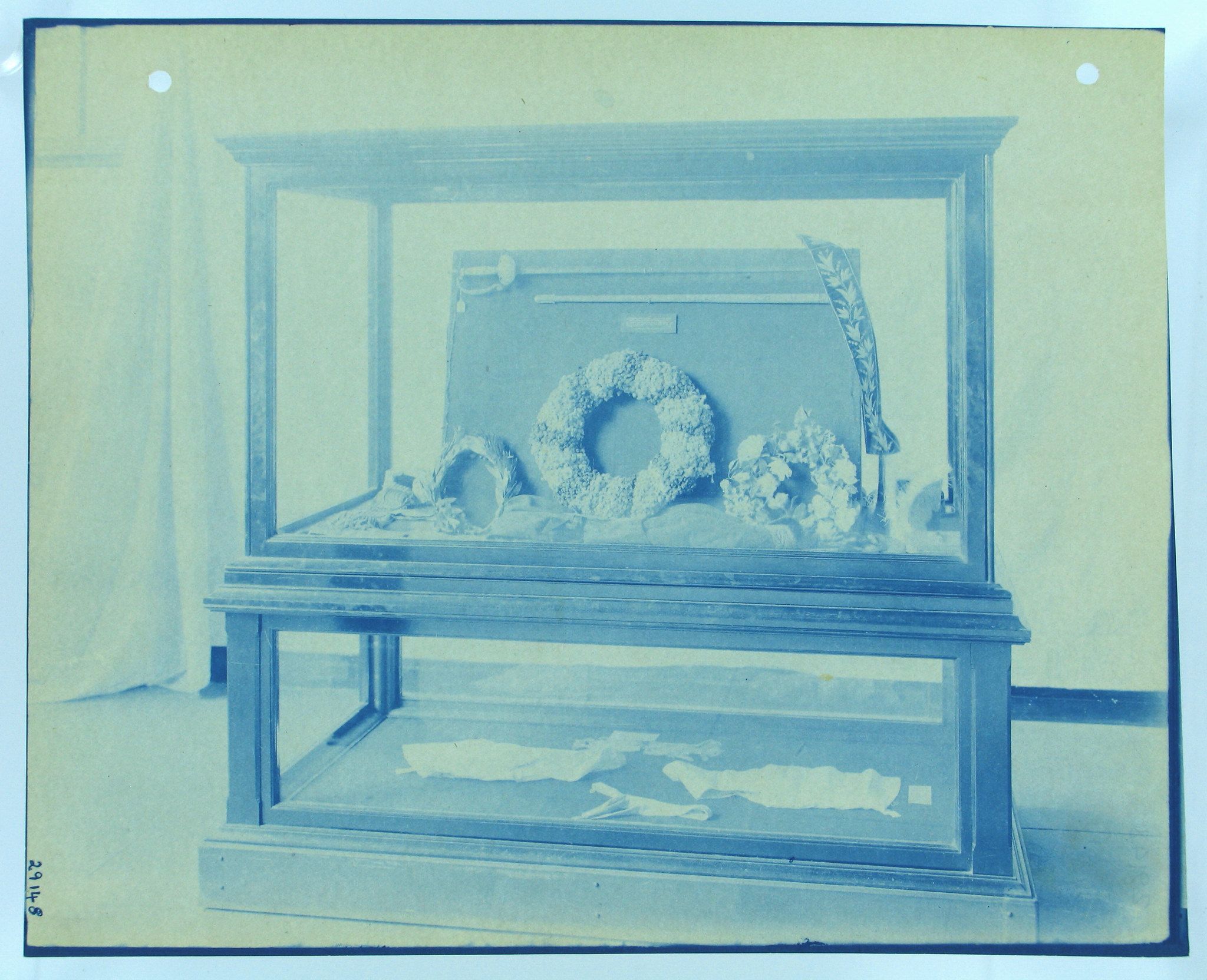 Vintage cyanotype photograph of a small coffin displayed inside a glass case, adorned with a wreath and flowers.