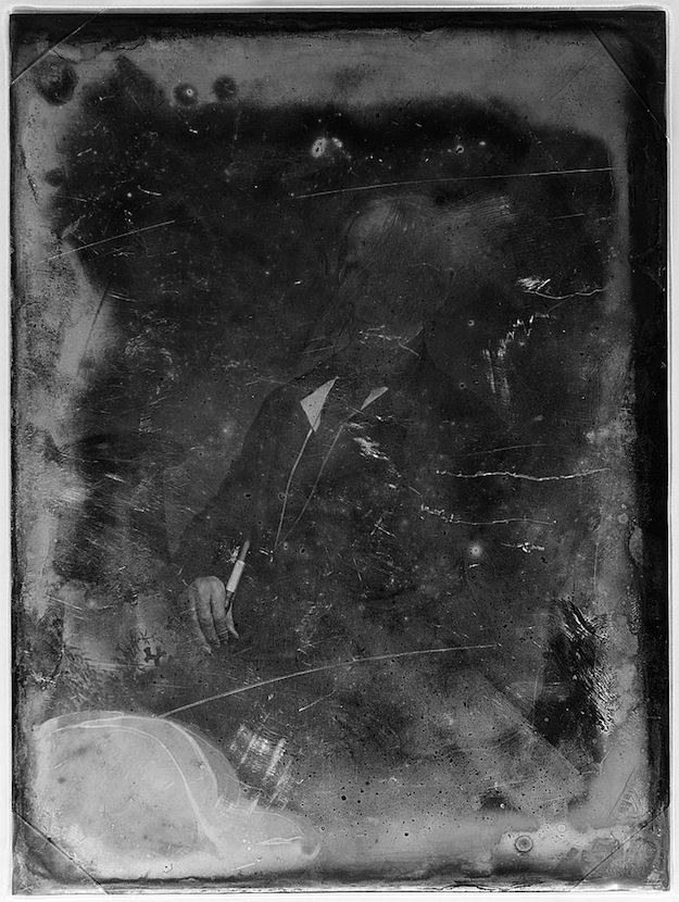 A vintage-style, black and white photograph of a person in a dark suit, holding a long, thin object. The image is heavily distressed with scratches, stains, and areas of emulsion loss.