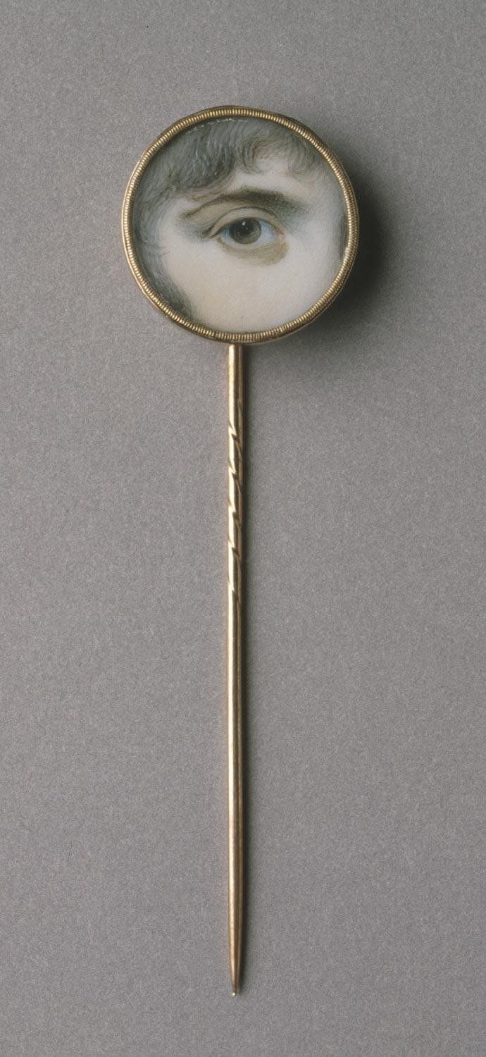 Close-up of a vintage brooch featuring a realistic depiction of a human eye within a circular gold frame, attached to a long, slender pin.