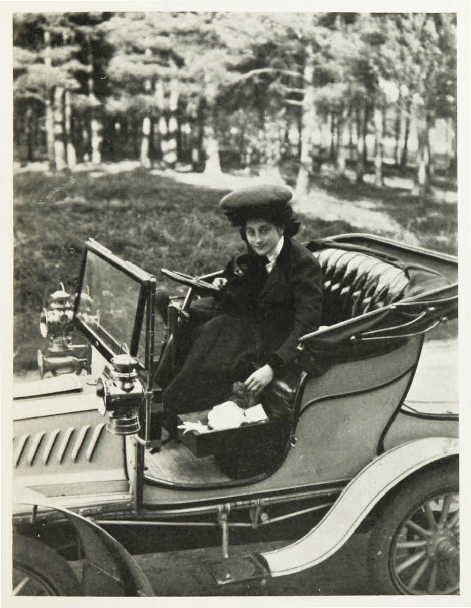 A vintage black and white photograph of a woman driving an early automobile.