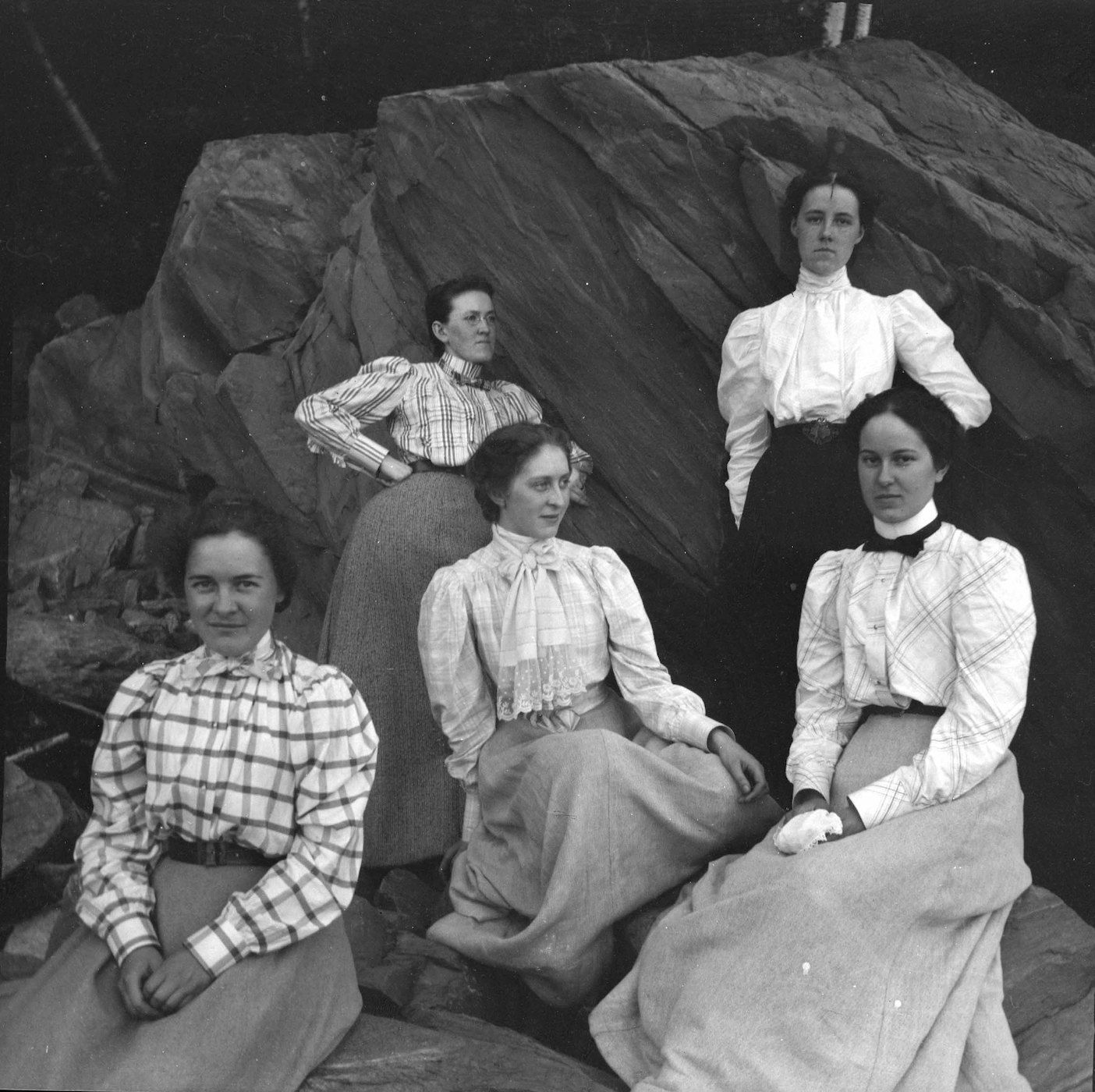 A black and white photograph of five women posing in front of a large rock formation. The women are dressed in clothing typical of the late 19th or early 20th century.