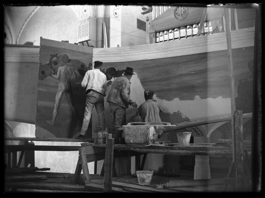 A black and white photograph of artists working on a large mural in a studio. Several men are positioned around a massive canvas or wall section, seemingly in the process of painting. They are standing on a complex scaffolding structure, which appears to be constructed from wooden planks and supports.