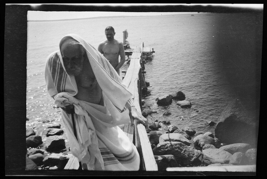 A black and white photograph of two men at a rocky shoreline. One man is ascending a wooden ladder from the water, draped in a light-colored cloth, while another man stands further up the ladder.