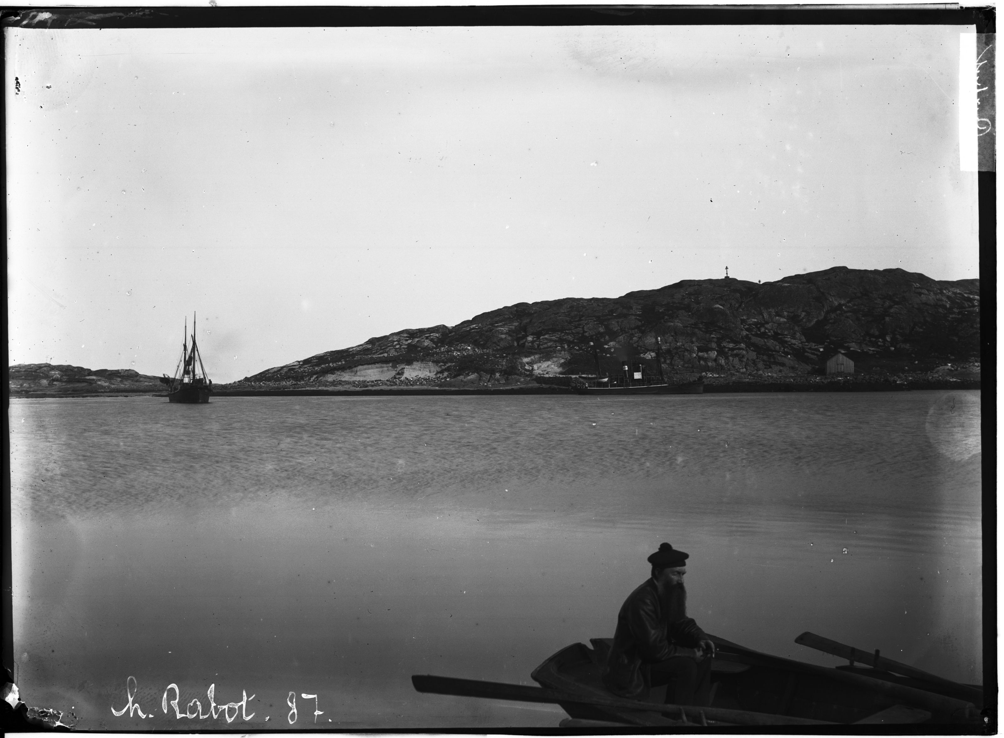 A vintage black and white photograph of a man in a boat on the water with a ship and hilly landscape in the background.