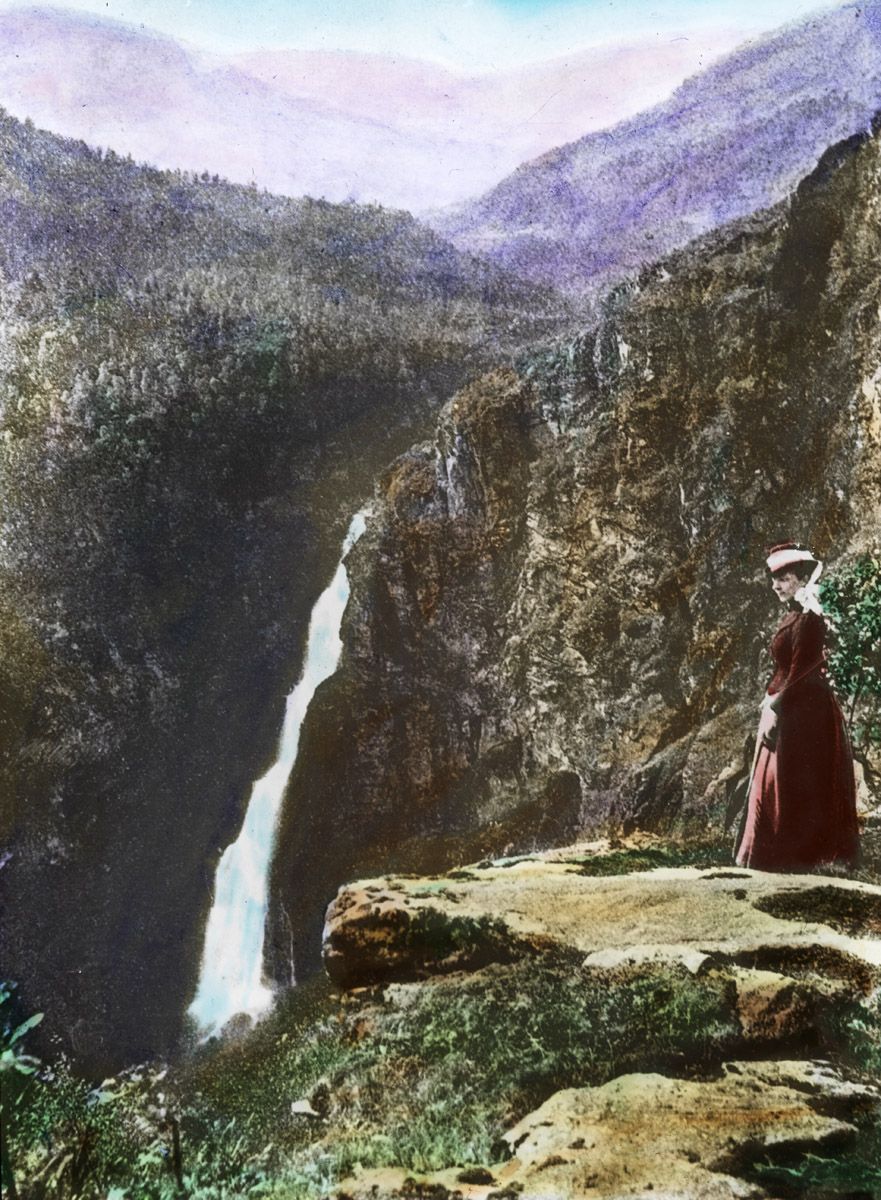 A vintage, hand-colored photograph of a woman standing on a rocky outcrop overlooking a large waterfall in a forested valley.