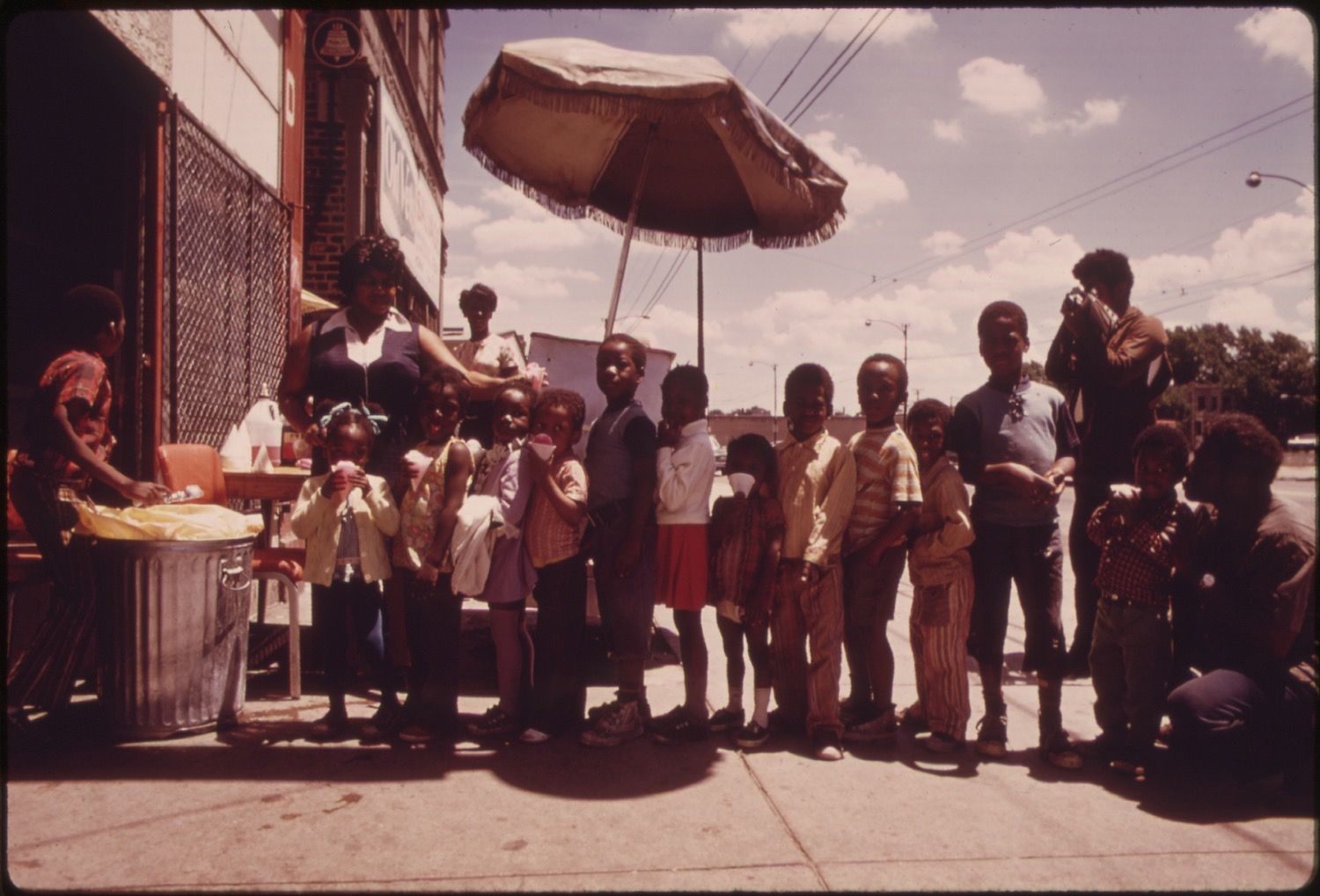 A group of children line up on a sidewalk, waiting for something from a man behind a table. Another man with a camera documents the scene in an urban setting.