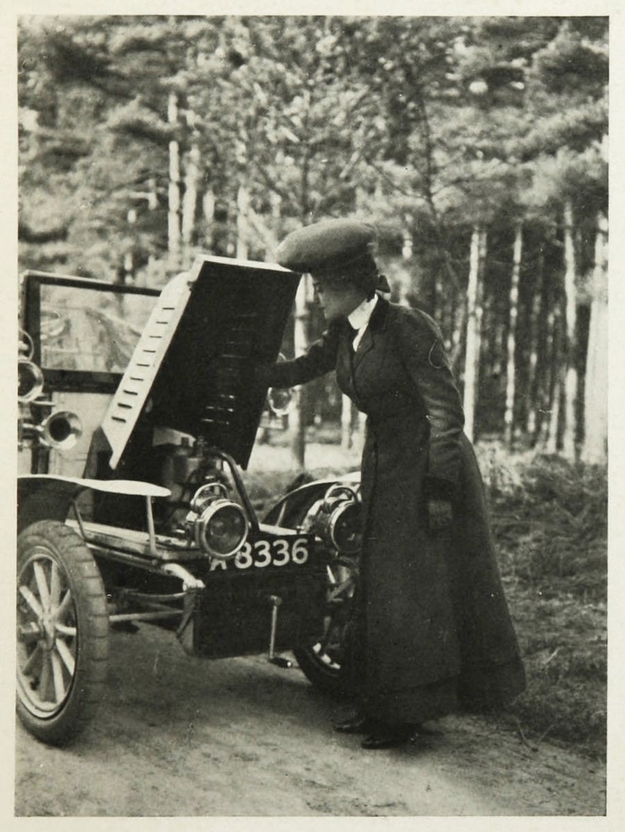A vintage black and white photograph of a woman in a long dress and hat inspecting the engine of an early automobile in a wooded area.