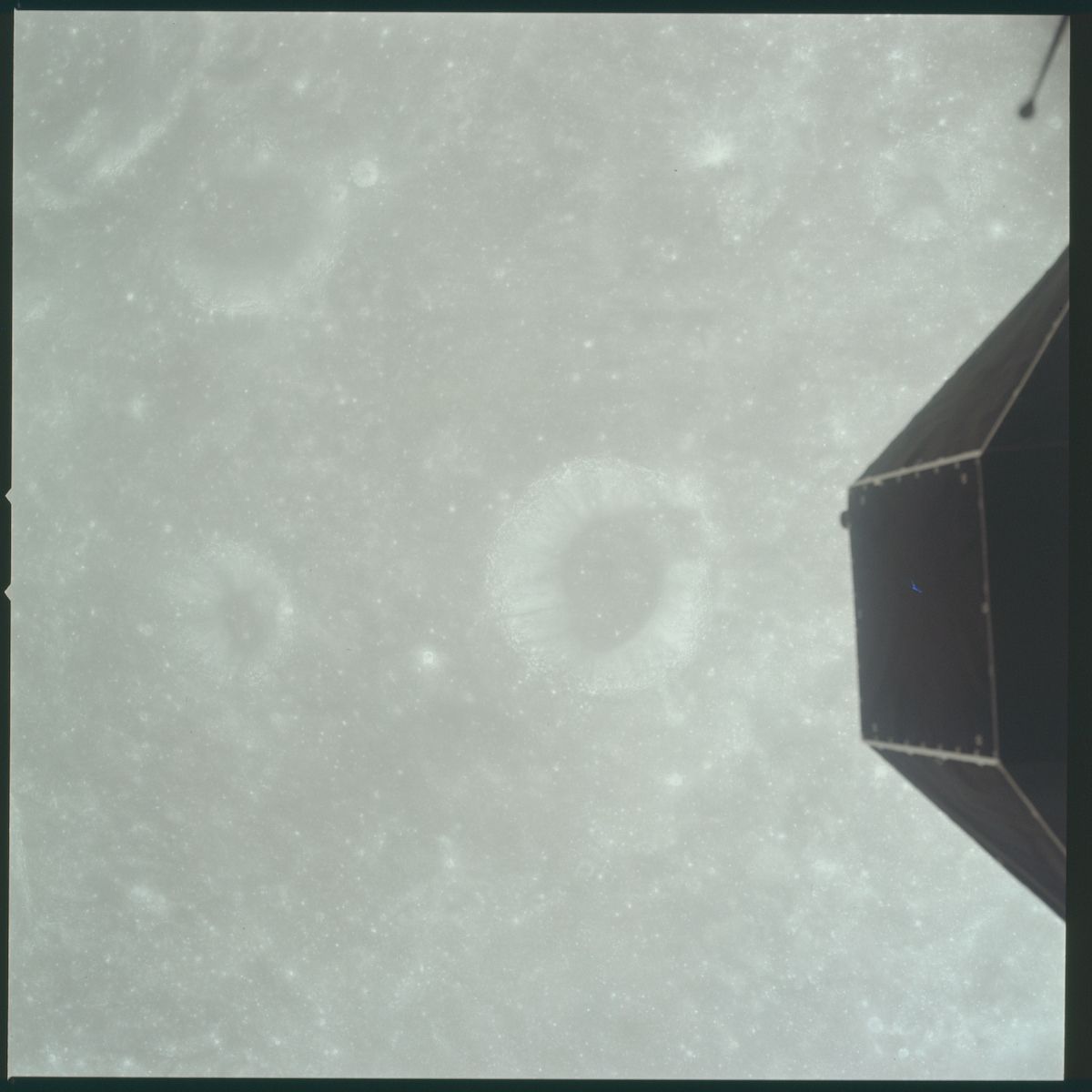 A grainy image of a pale green planet with a faint ring system, taken from a spacecraft with a hexagonal structure partially visible in the frame.