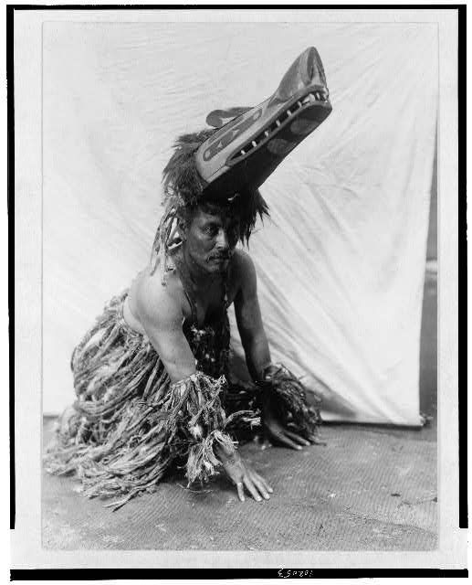 A black and white photograph of a person wearing a wolf mask and feathered costume, crouched on the floor in a dramatic pose.