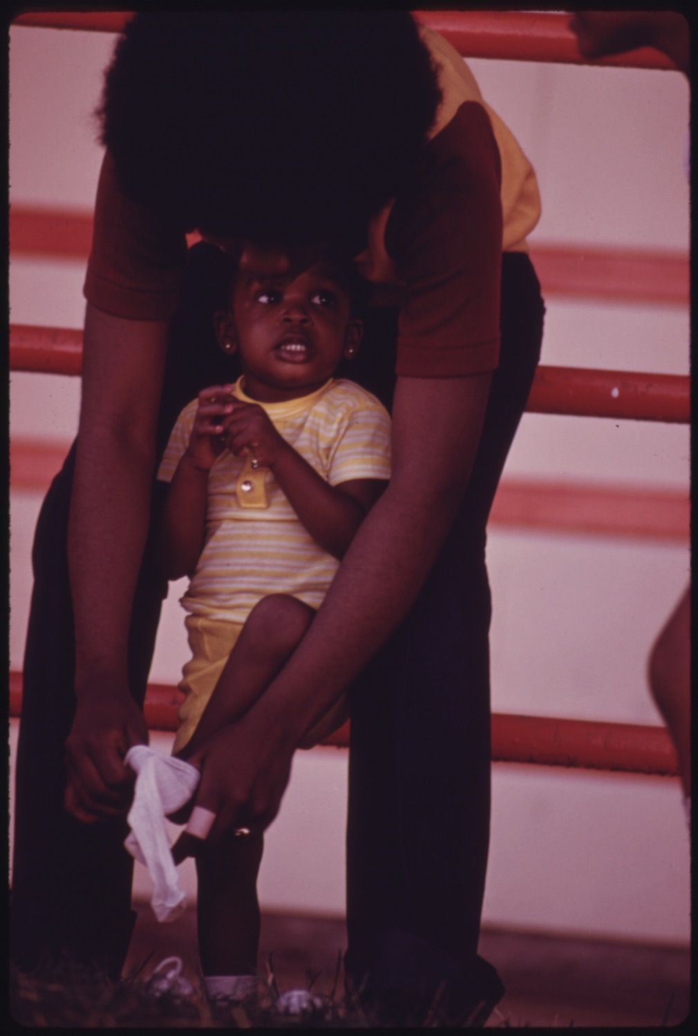 Close-up of a young Black child being held by an adult, wearing a yellow and white striped shirt and light-colored pants, with a simple wall as the background.