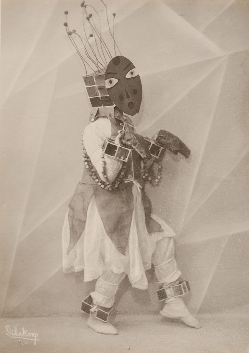 A black and white photograph of a person in a geometric mask and costume, with wire-like headgear, standing against a geometric background. The photograph is signed 'Duboiskoop 1920'.