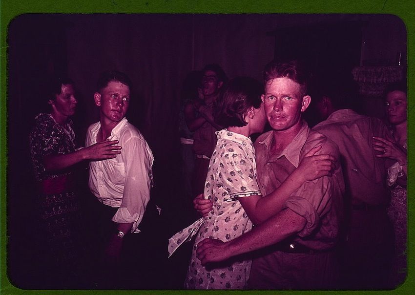 A color photograph of a soldier embracing a woman in a crowded, dimly lit space. The scene is dominated by a magenta/purple color cast, and the atmosphere is lively and energetic.