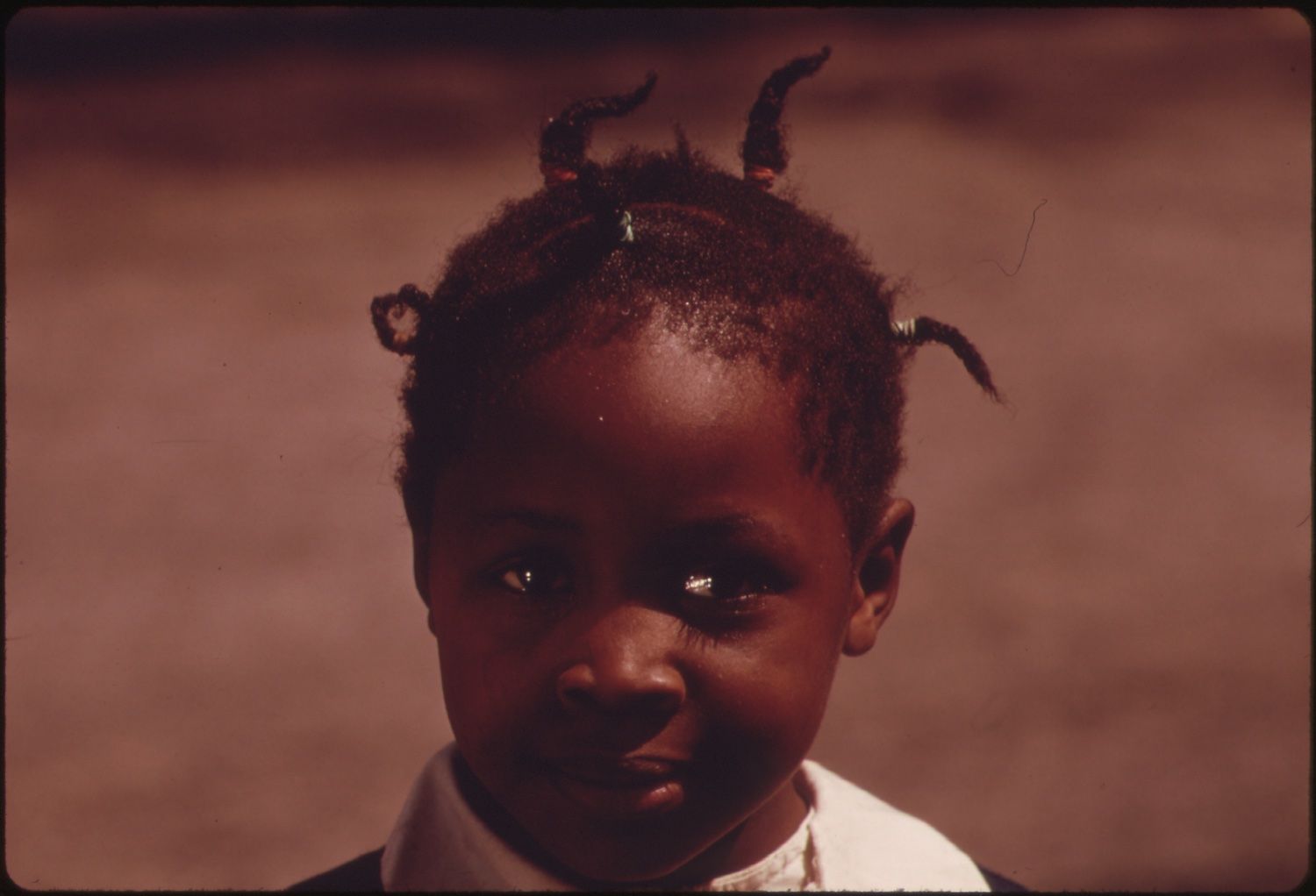 Close-up portrait of a young child with dark skin and beaded hair, looking directly at the camera with a serious expression.
