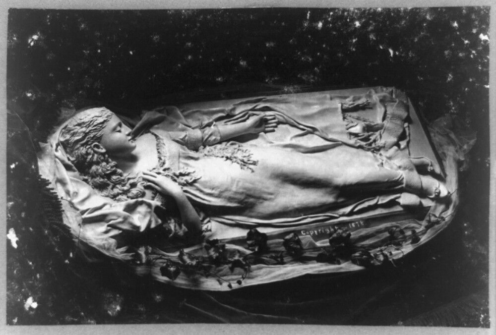 A black and white photograph of a person lying in a coffin or bed, decorated with a floral arrangement.