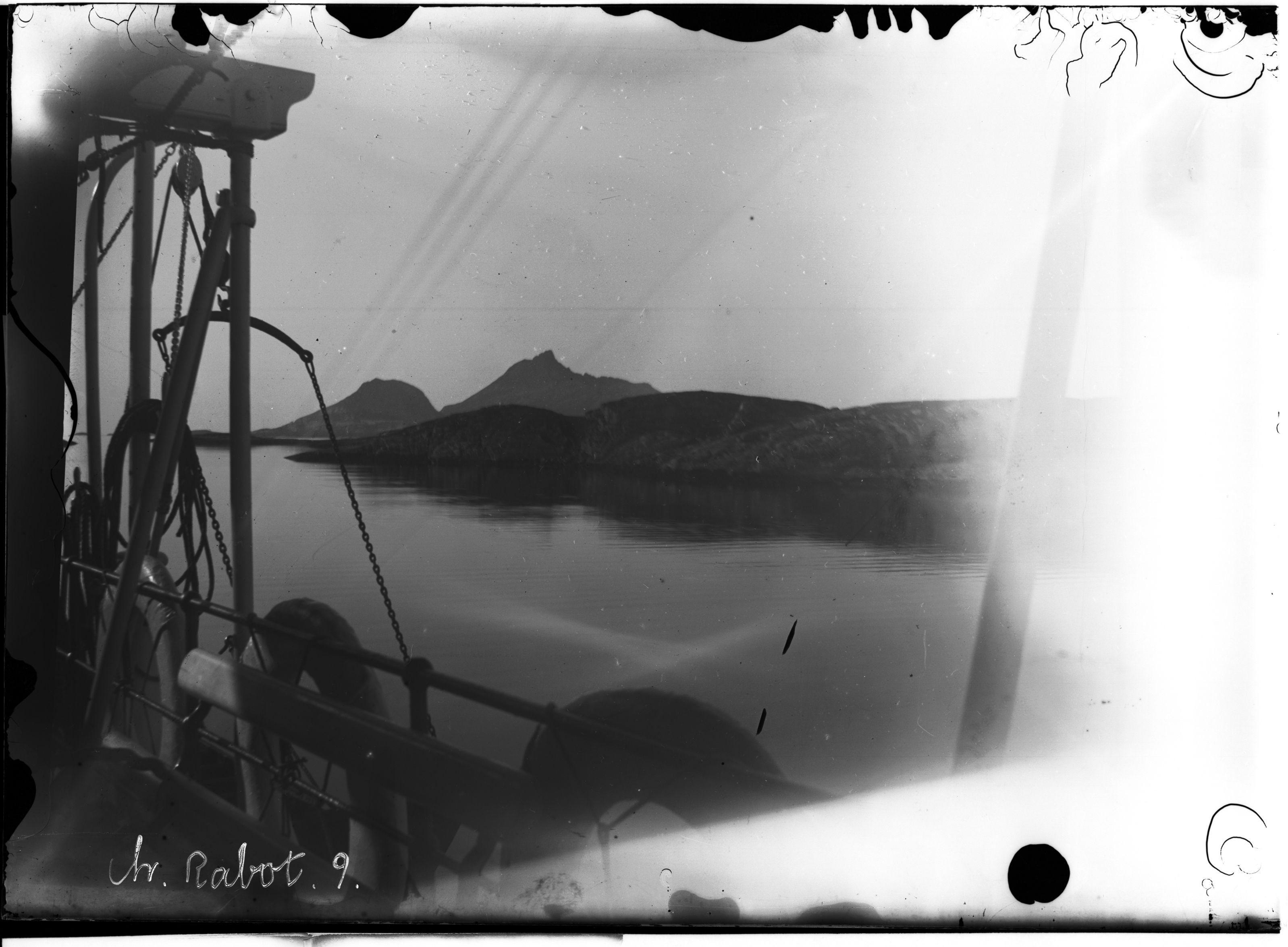 A vintage black and white photograph taken from the deck of a ship, showing mountains in the distance and ship rigging in the foreground.