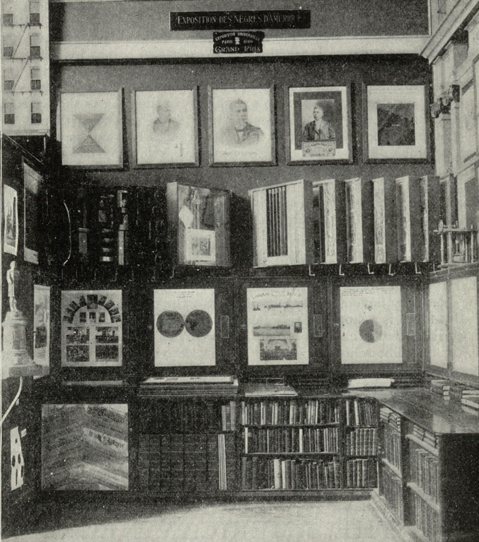 Black and white photograph of an exhibition room with walls covered in framed artwork and shelves filled with books.