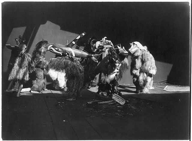 A black and white photograph of a group of performers wearing elaborate, full-body masks and costumes on a stage.