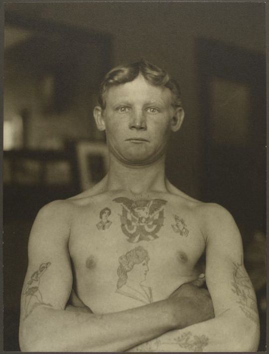 A sepia-toned portrait of a young man with extensive tattoos covering his upper body.