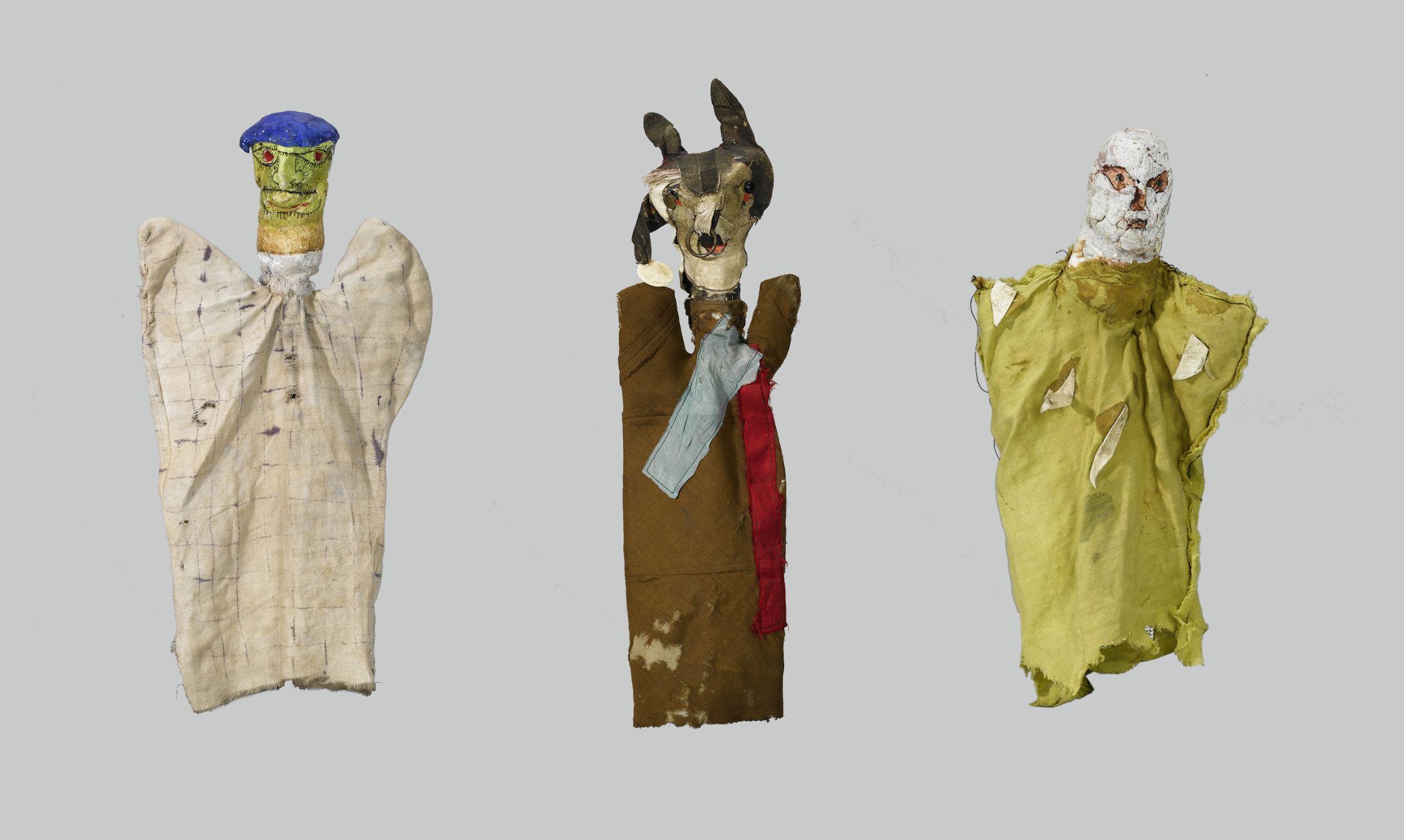 Three handmade puppets against a light gray background. Each puppet is constructed with a roughly formed head and a draped, fabric body, giving them a primitive, folk-art aesthetic.