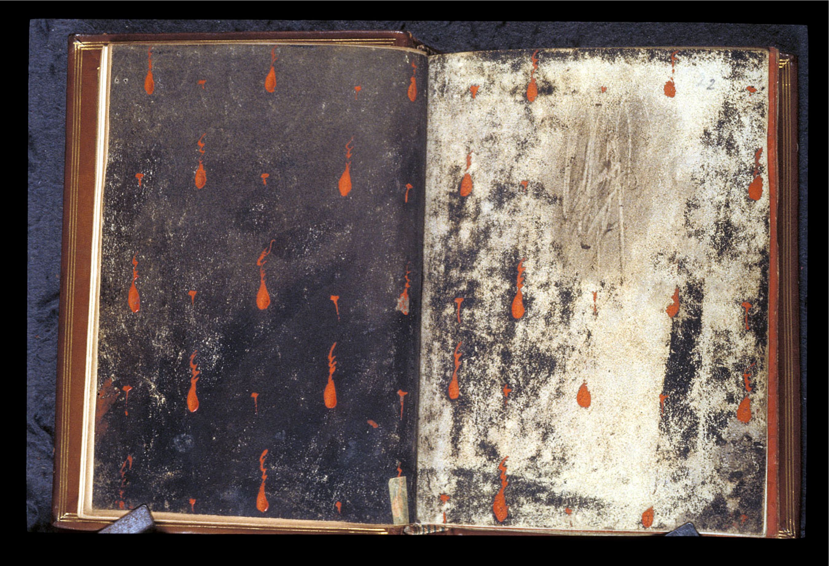 An open, aged book with dark, speckled pages and orange teardrop markings.