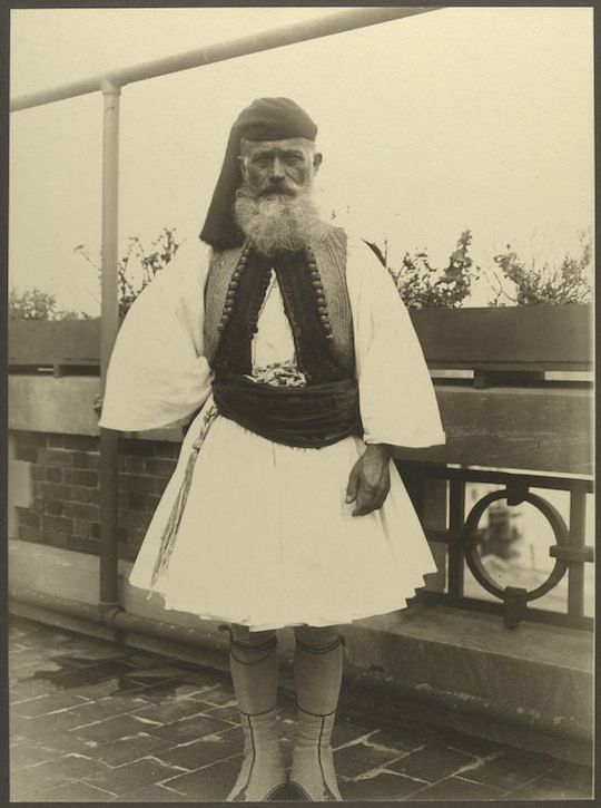 Sepia-toned photograph of a man in traditional Albanian attire standing on a balcony.
