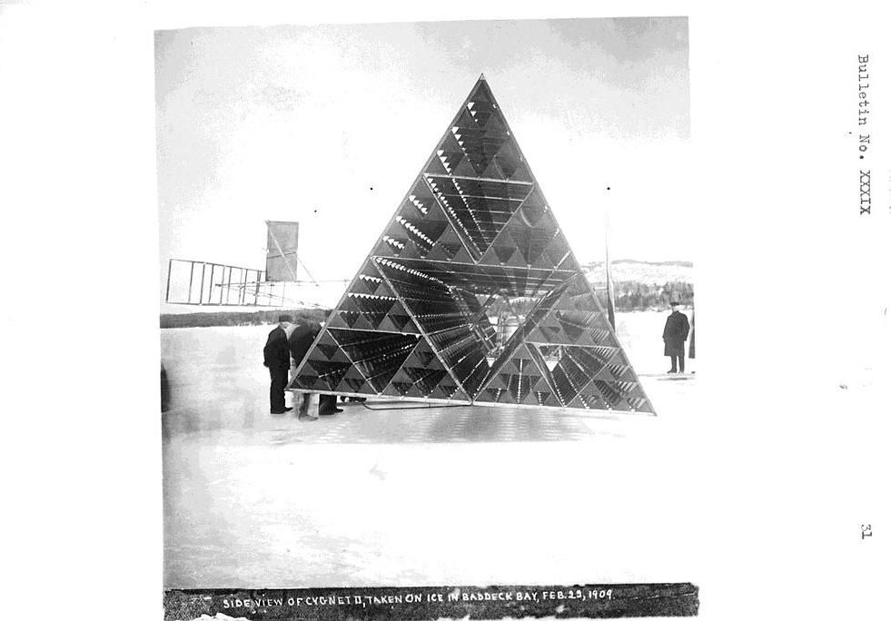 A vintage black and white photograph of a large ice pyramid constructed on a frozen bay in Baddeck Bay on February 29, 1904. Several people are gathered around the base of the pyramid, and a flag is visible in the background.