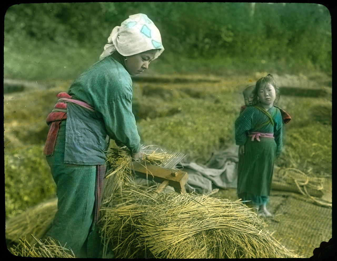 A historical image of two people working in a field, one adult and one child, both dressed in traditional clothing, with a haystack in the foreground and a field of golden grass and trees in the background.