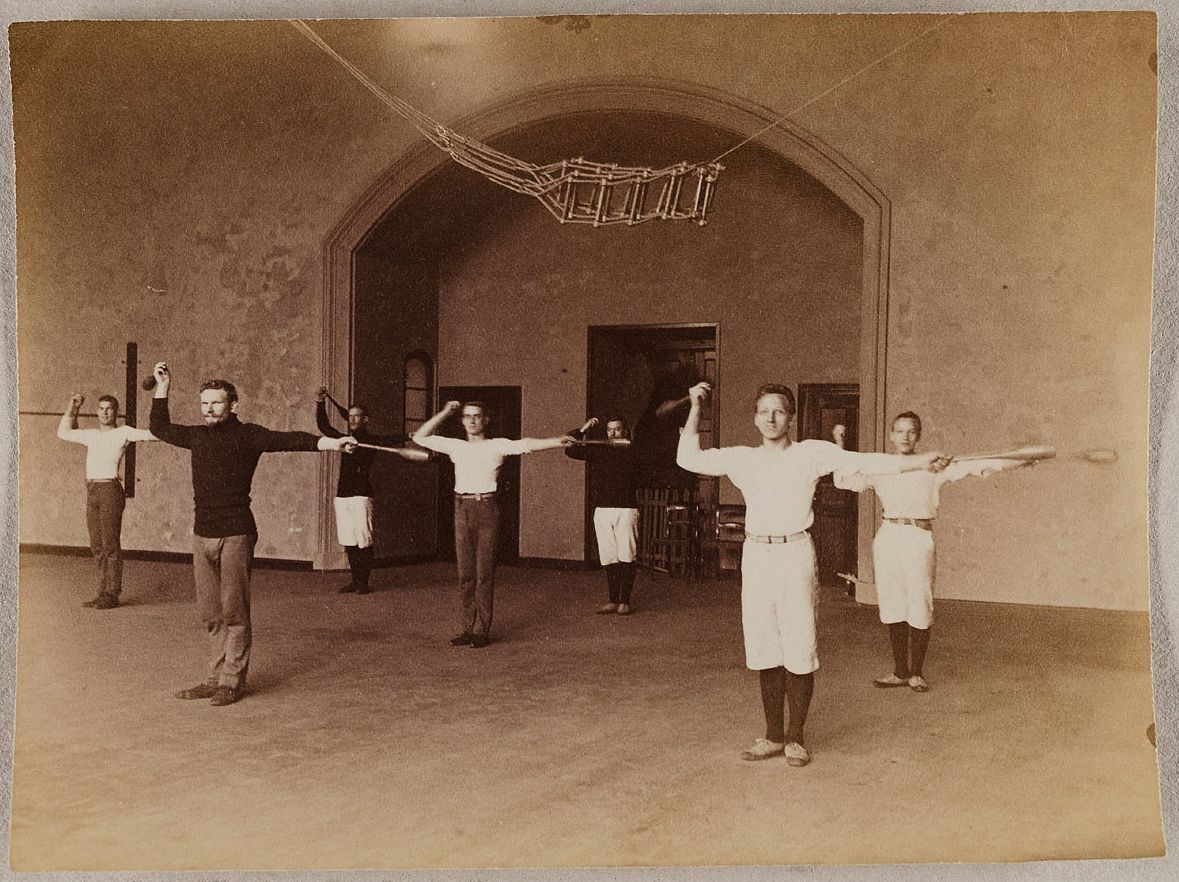 A vintage photograph of six men in a gymnasium performing a synchronized exercise with poles.