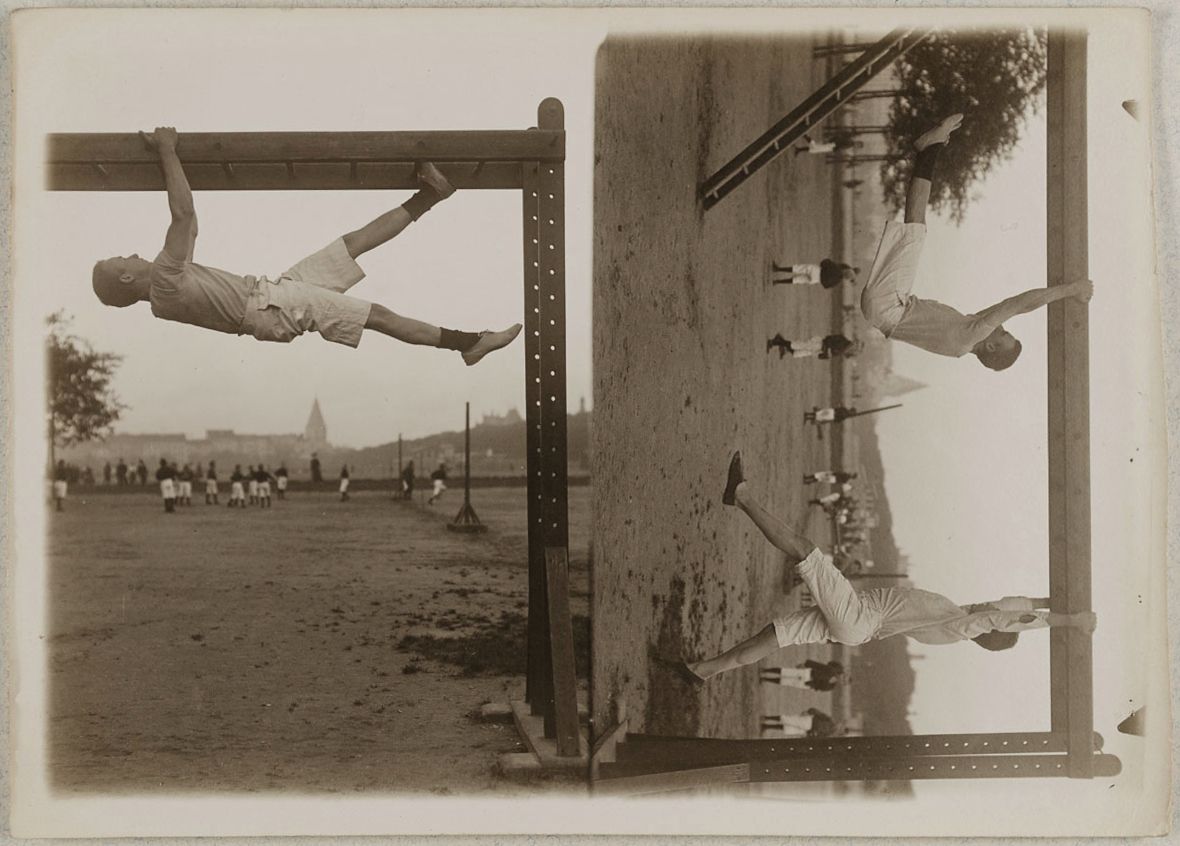 A sepia-toned photograph of boys doing gymnastic exercises on a wooden structure in a park setting.