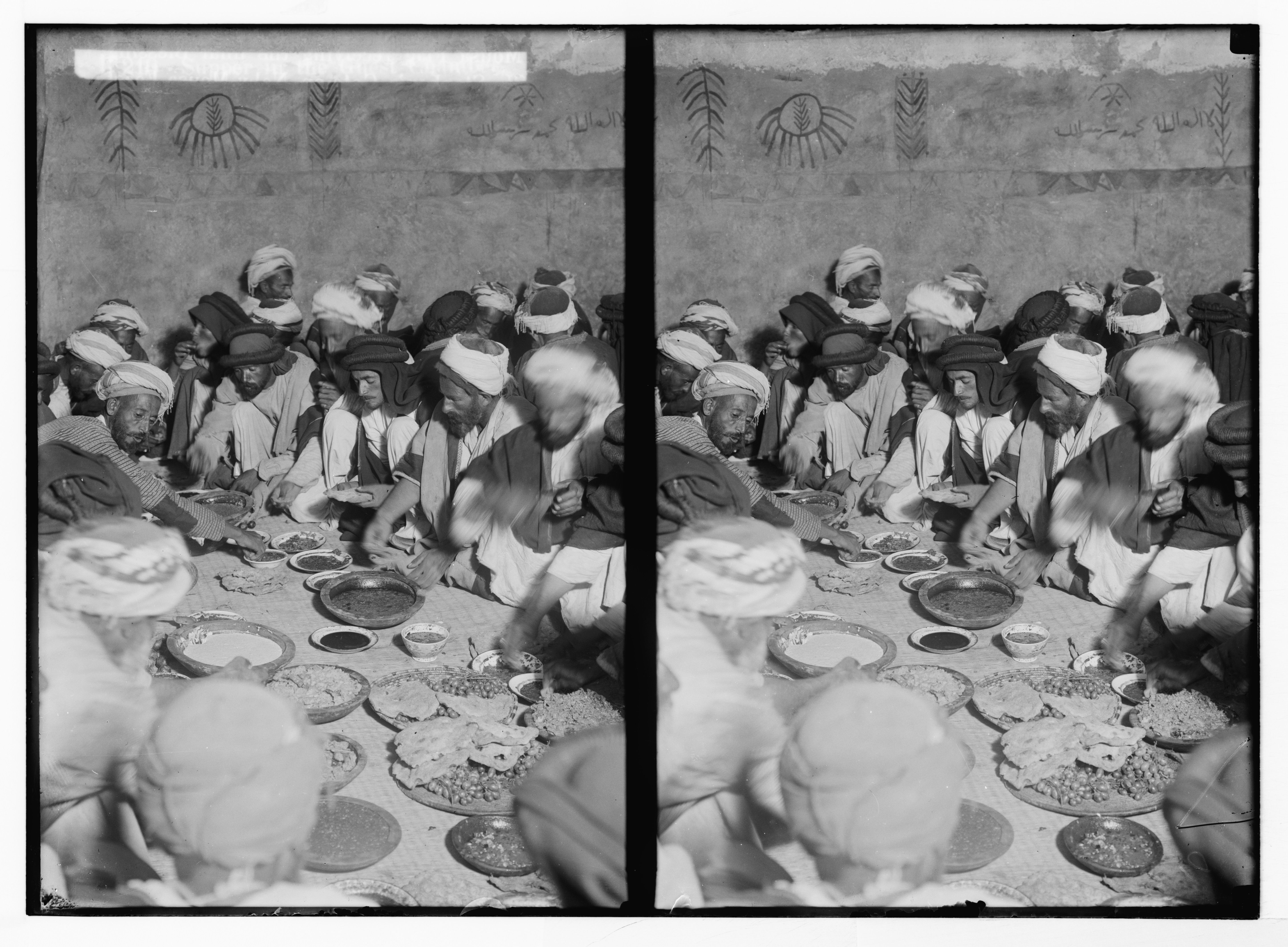 Stereoscopic photograph of a group of men seated around a table laden with food, dressed in traditional clothing, with a decorated wall in the background.