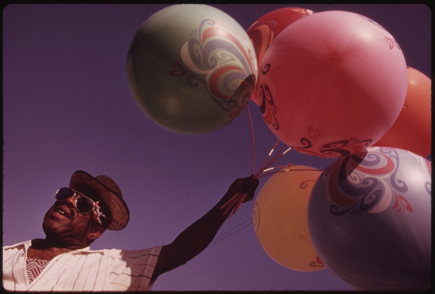 Low-angle view of a man holding a bunch of colorful, patterned balloons against a purple sky.