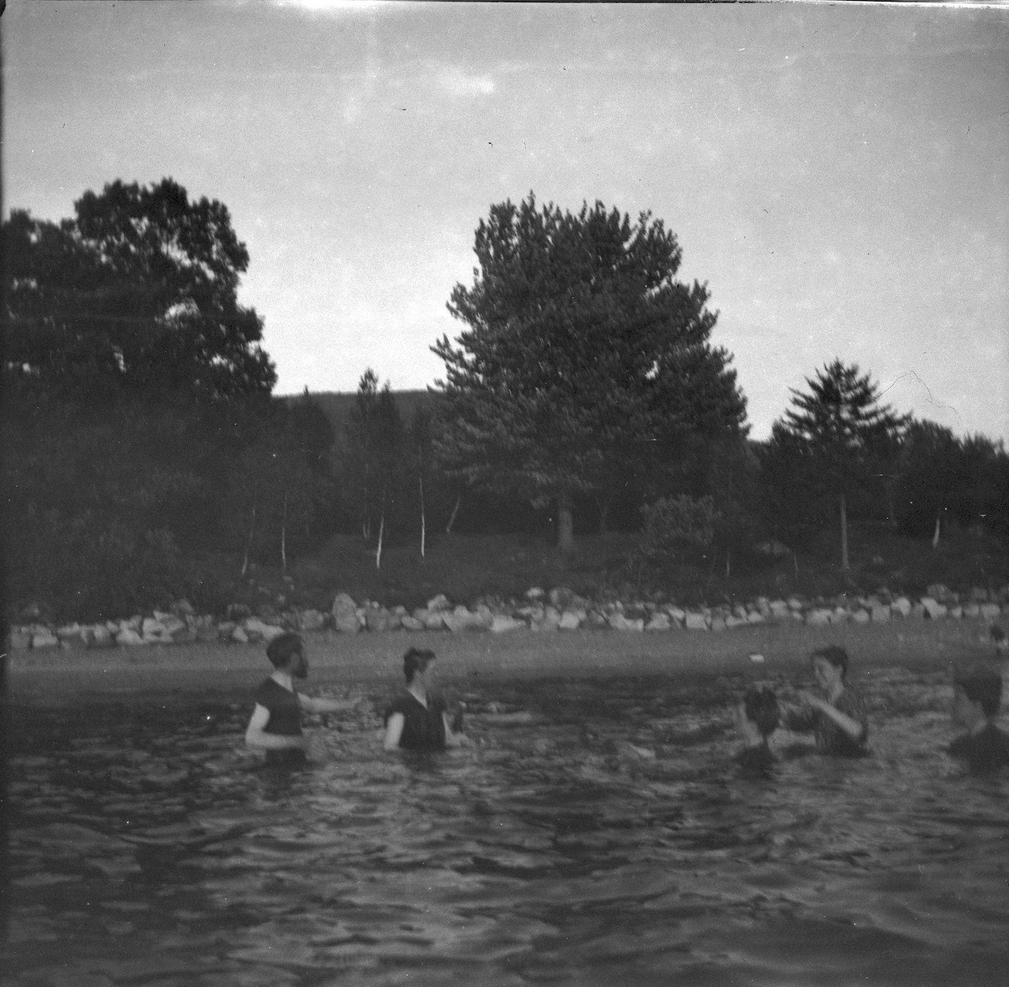 A vintage black and white photograph of people swimming and relaxing on a crowded beach, with trees in the background and an overcast sky.
