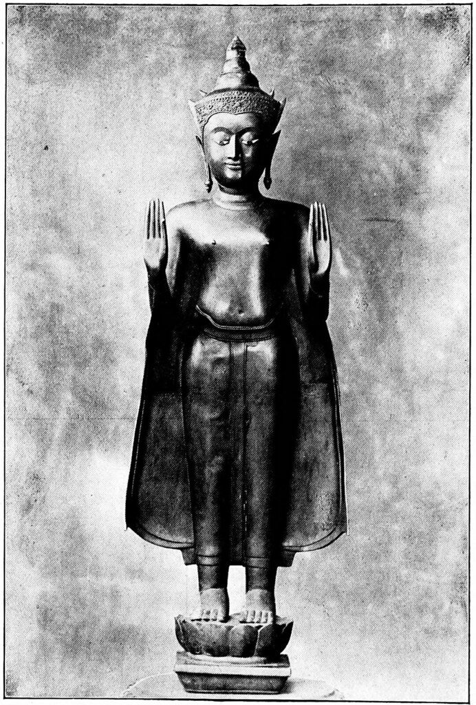 Black and white photograph of a standing Buddha statue with hands raised in a gesture of blessing, standing on a lotus-shaped pedestal.