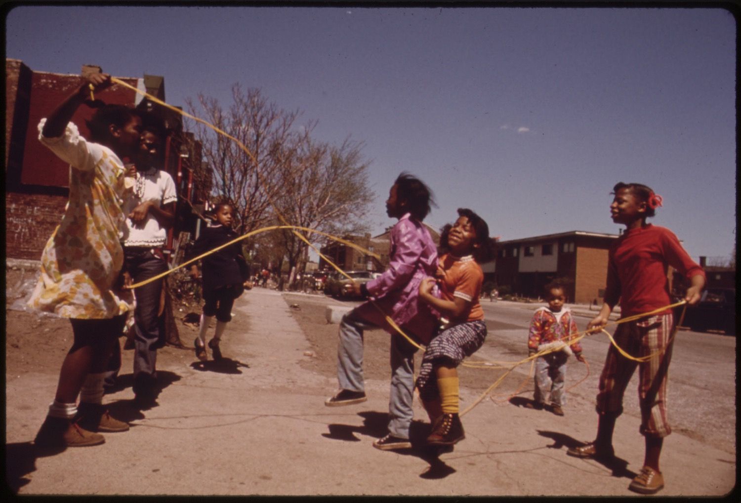 A group of children playing double dutch jump rope on a sunny street in a residential neighborhood.