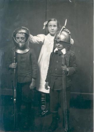A vintage black and white photograph of three children dressed in theatrical costumes, including suits of armor and swords.