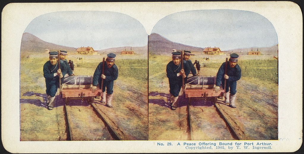 Stereoscopic photograph of soldiers pulling a machine across a field, with buildings and hills in the background. Text below reads: “No. 29. A Peace Offering Bound for Port Arthur. Copyrighted, 1905, by T. W. Ingersoll.”
