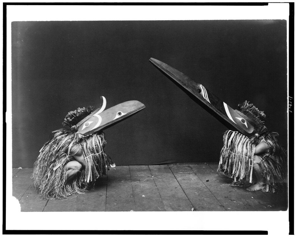 Two figures wearing elaborate bird-like masks, seated on a wooden floor against a black background.