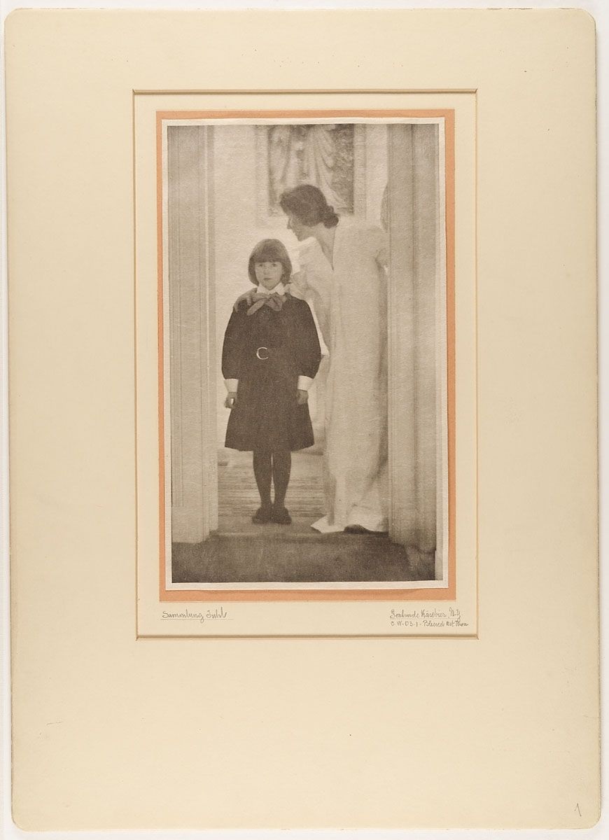 A vintage black and white photograph of a woman and a child in an interior room, mounted on a cream-colored mat.