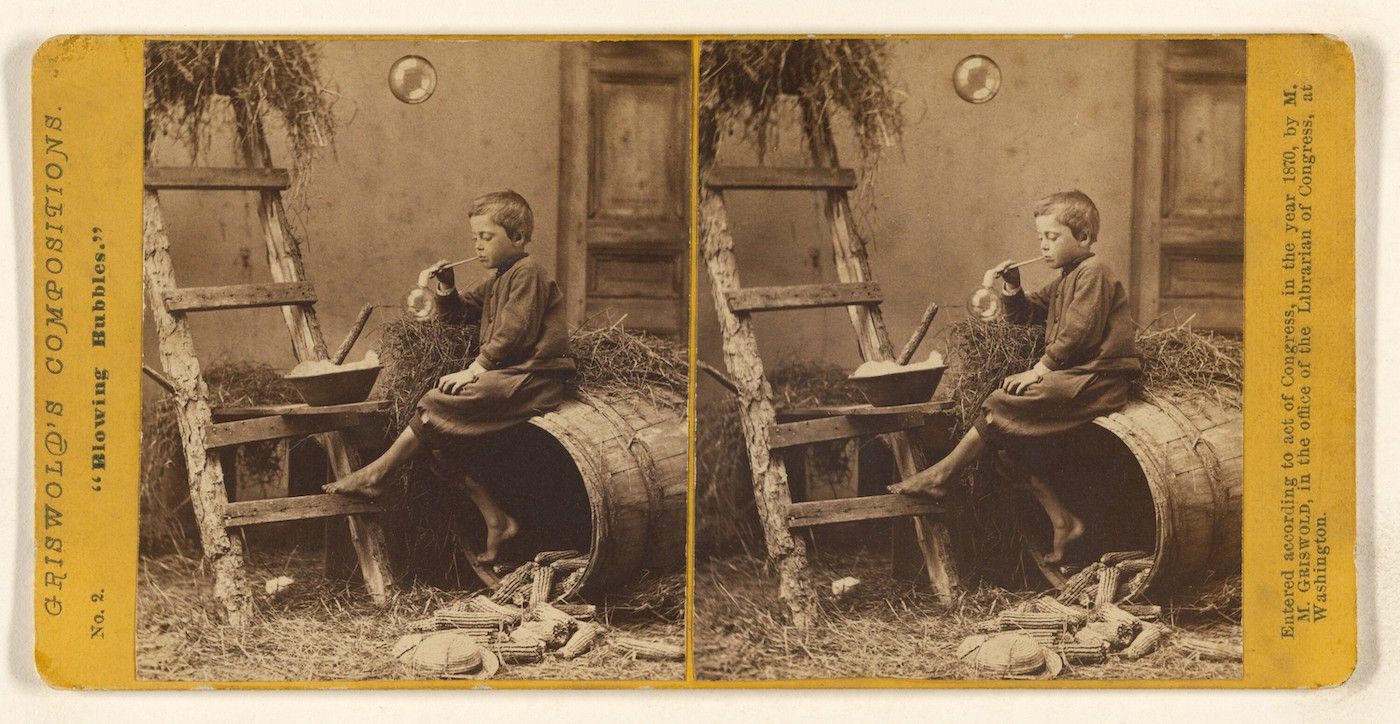 Stereoscopic photograph of a young boy blowing bubbles while sitting on a barrel in a rustic outdoor setting.