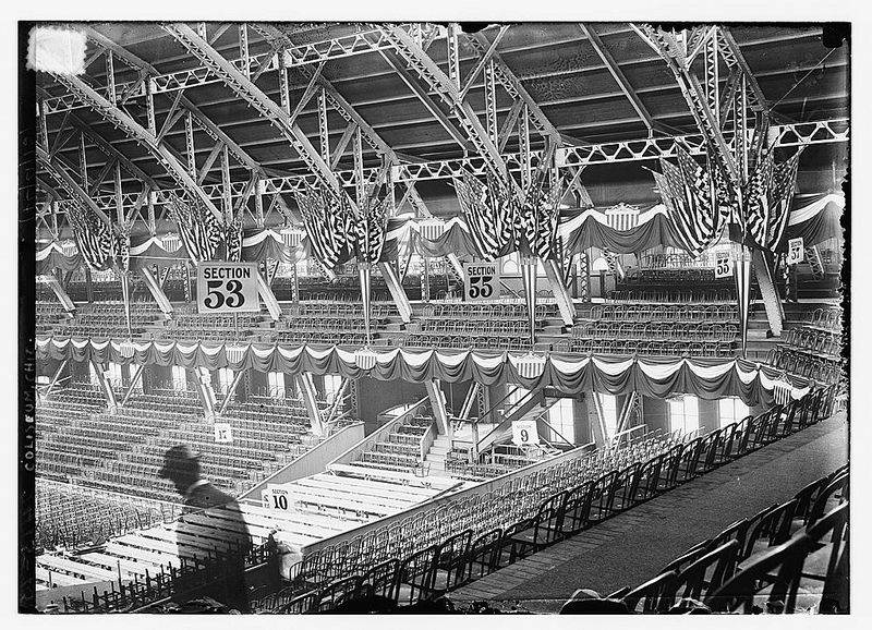Black and white photograph of the interior of a large indoor arena with tiered seating, section numbers, flags, and a stage.