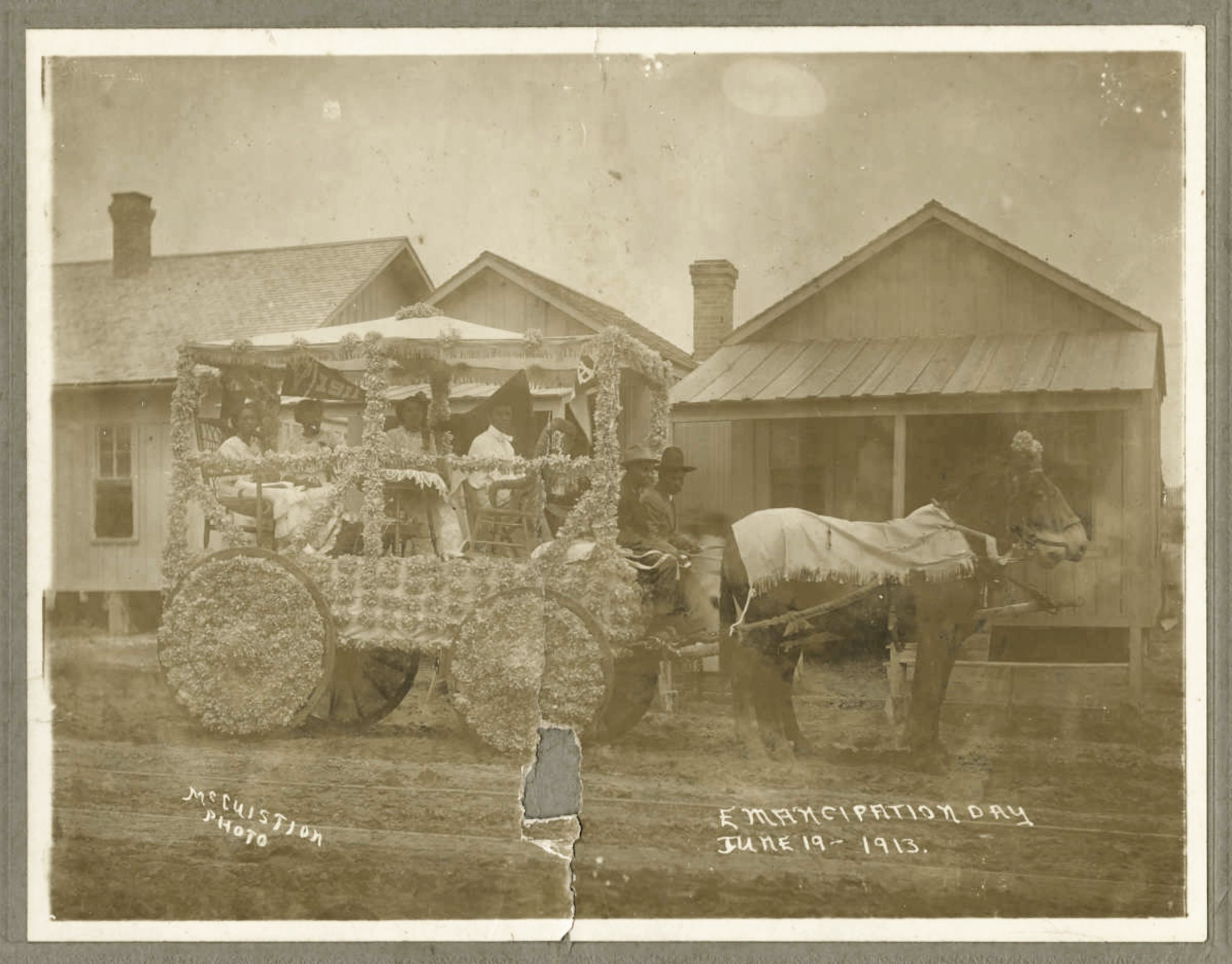 A sepia-toned photograph from June 19, 1913, commemorating Emancipation Day. The central focus is a horse-drawn carriage, heavily decorated with what appears to be flowers or foliage. Two horses are harnessed to the carriage, standing in a grassy area. Behind the carriage, there are two single-story buildings with gabled roofs and visible windows.