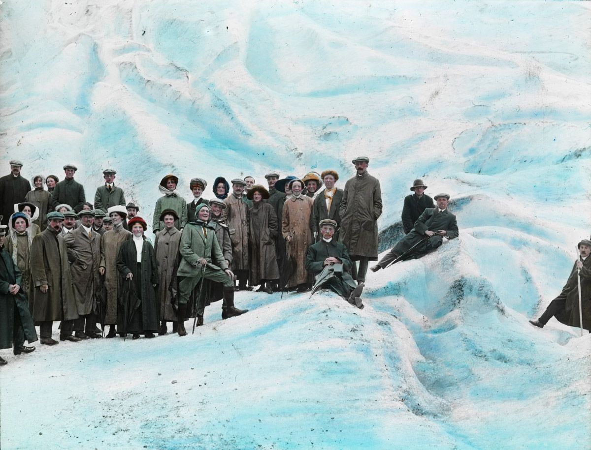 A vintage, colorized photograph of a large group of people standing on a snowy, mountainous landscape.