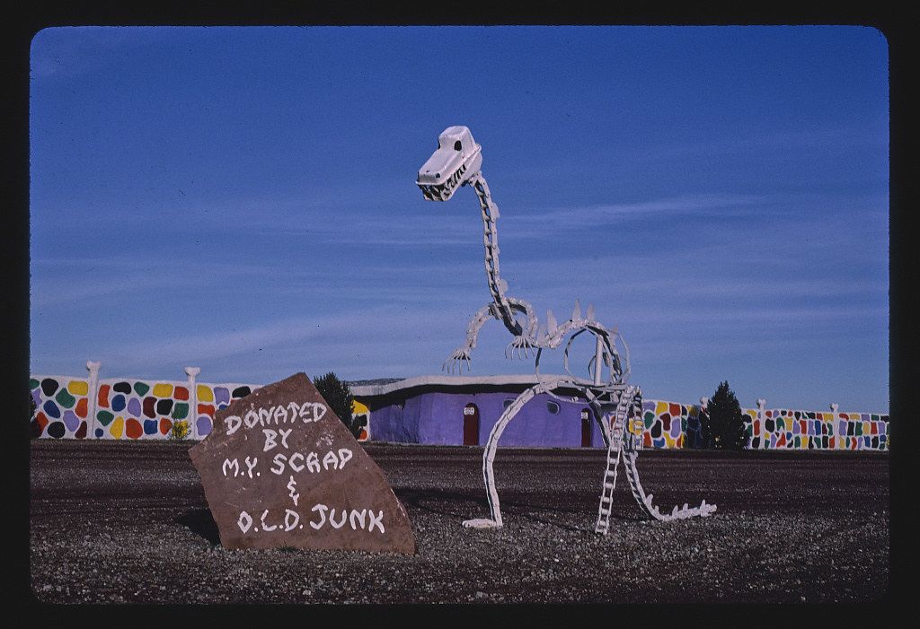 A large, skeletal dinosaur sculpture stands in front of a low, flat building under a deep blue sky. The dinosaur is constructed from metal and appears to be made of scrap parts. In front of the dinosaur is a rough, triangular sign that reads "DONATED BY M.Y. SCRAP & O.C.D. JUNK" in painted lettering. The building behind the dinosaur is a single-story structure with a dark roof and a purple-colored wall. On either side of the dinosaur and building, there's a colorful fence made of what appears to be glass or plastic pieces embedded in a frame. The ground in front of the dinosaur and building is a dry, grassy field.