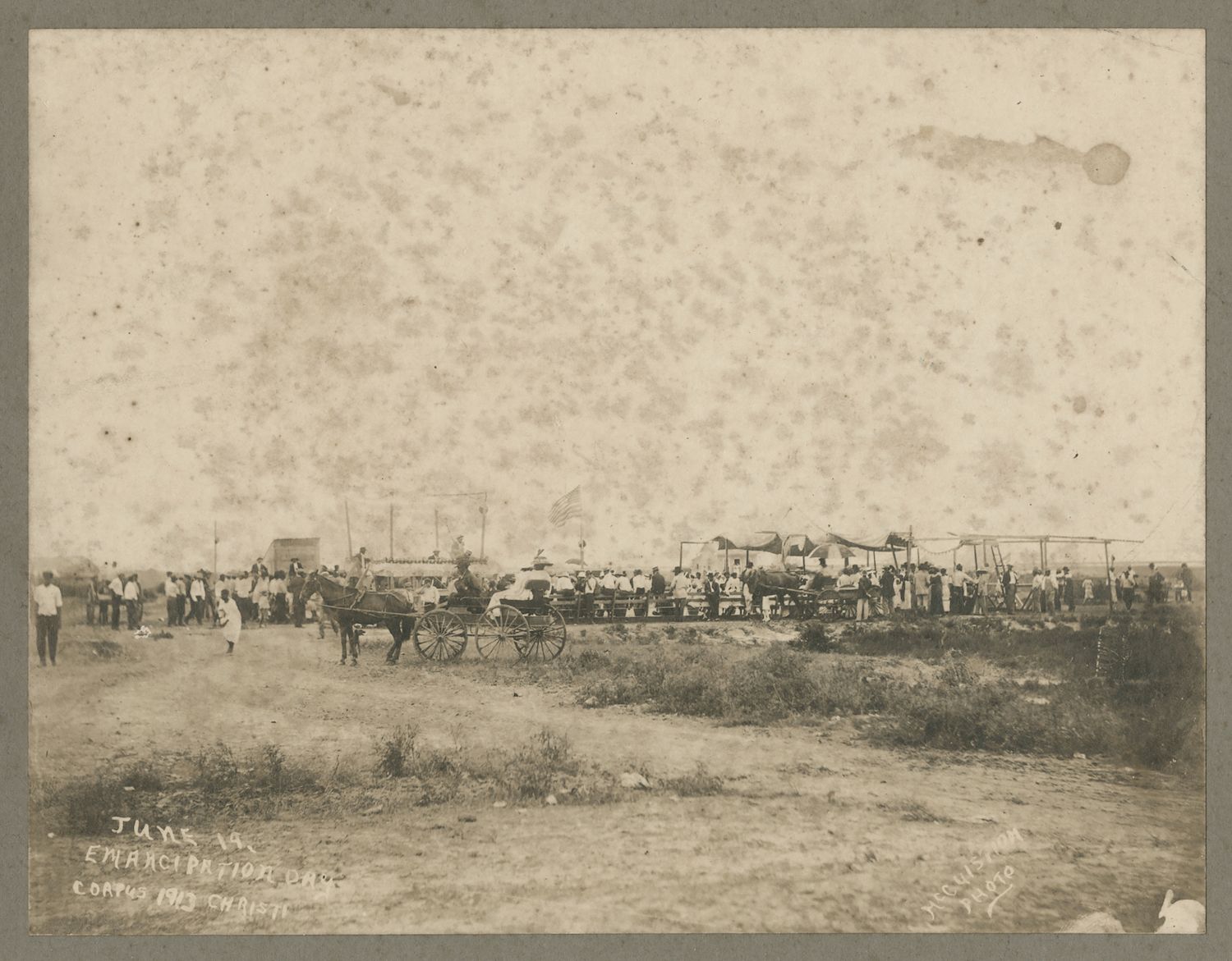 A sepia-toned photograph of a large outdoor gathering, likely a celebration or event, with a crowd of people, a horse-drawn carriage, flags, and tents in a field.