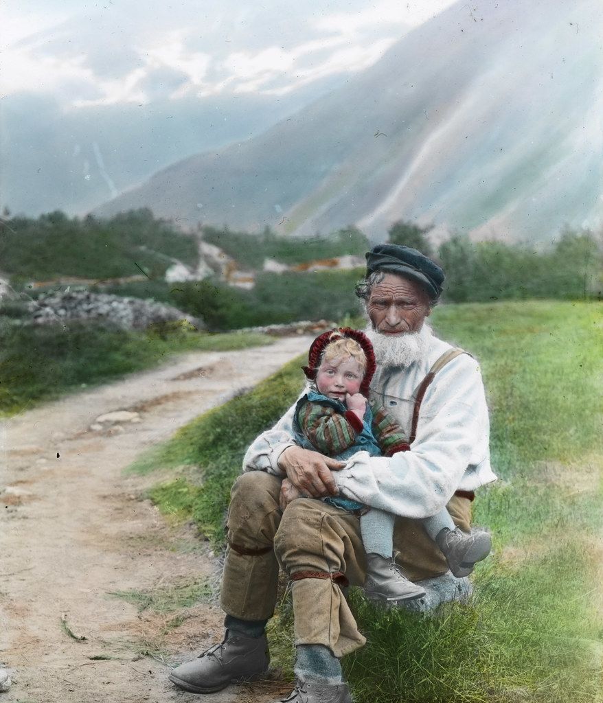 A vintage colorized photograph of an elderly man with a long white beard holding a young child in his lap on a grassy hill, with a dirt road and mountains in the background.