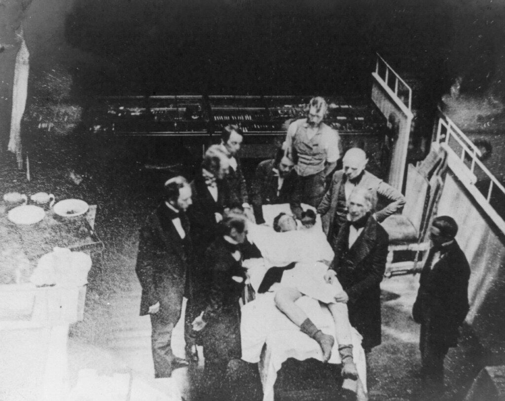 A black and white photograph of a group of men in suits surrounding a person lying on a bed, likely in a hospital setting.