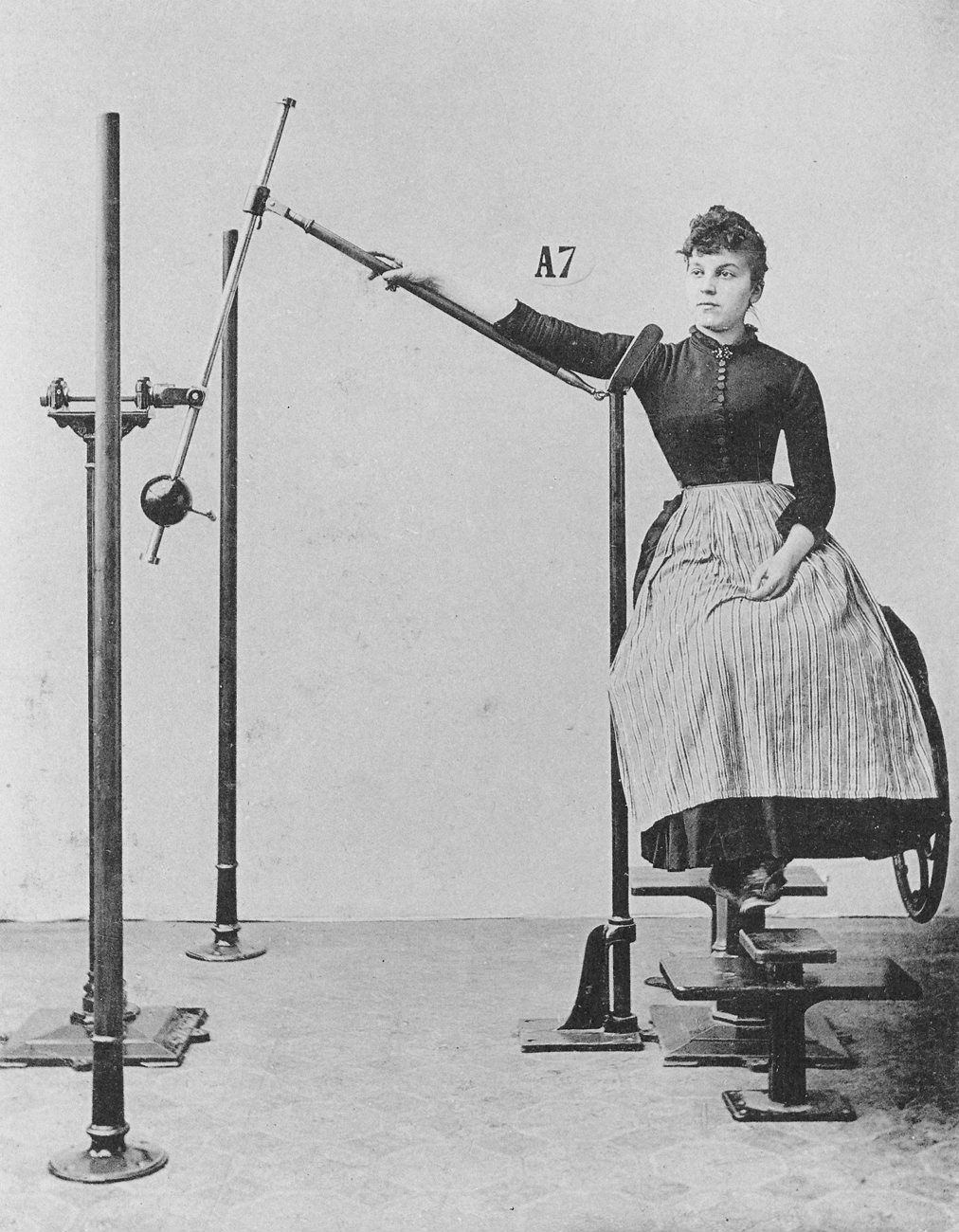 A vintage black and white photograph of a woman seated in a complex mechanical device, likely a scientific or medical instrument.