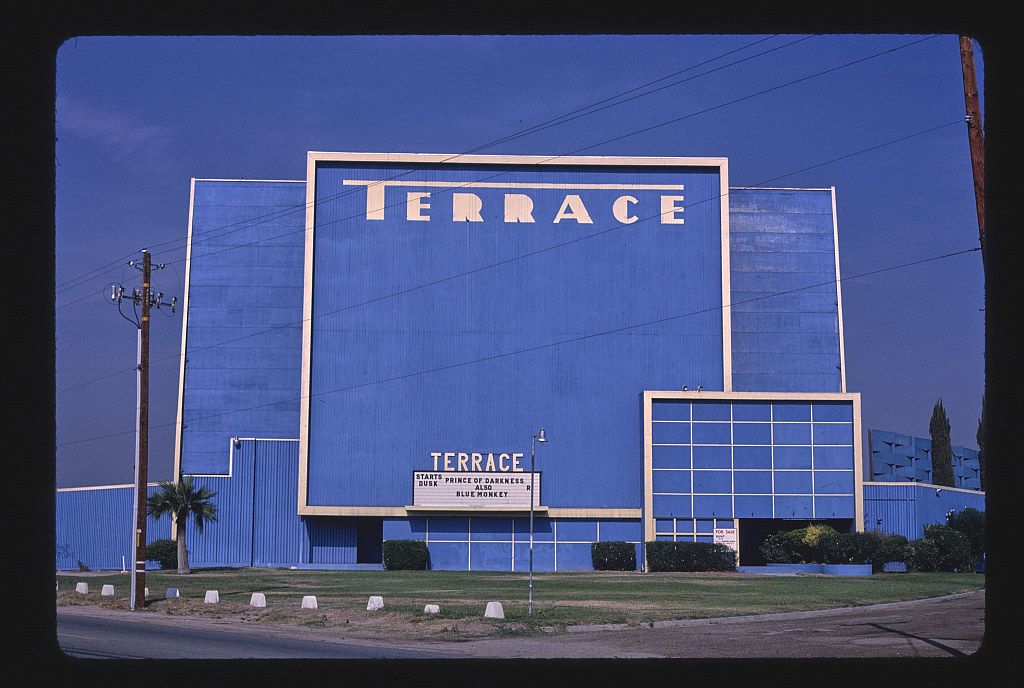 A bright blue building with the word "TERRACE" in large white letters, advertising the movies "Prince of Darkness" and "Blue Monkey". The building is set back from a road with a small patch of grass and a sidewalk in front. The sky is clear and blue.
