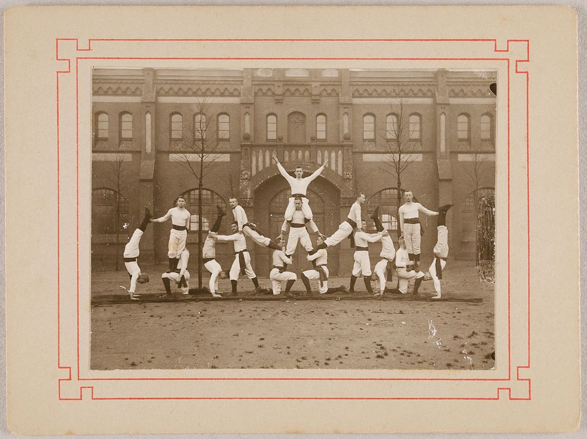 A vintage photograph of a group of men in athletic uniforms posing in front of a large brick building.
