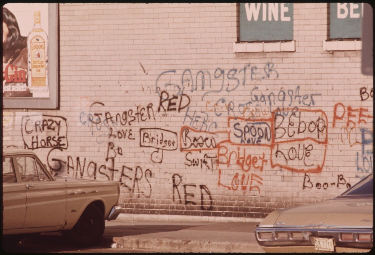A brick wall covered in colorful graffiti with a vintage car parked in front of it. A window with a sign for wine and beer is above the graffiti, and a poster is visible to the left.